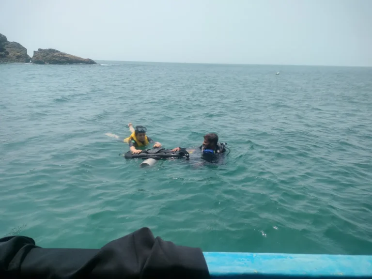 Two people in the water off the boat, one in a yellow vest and one in scuba gear, near rocky islets
