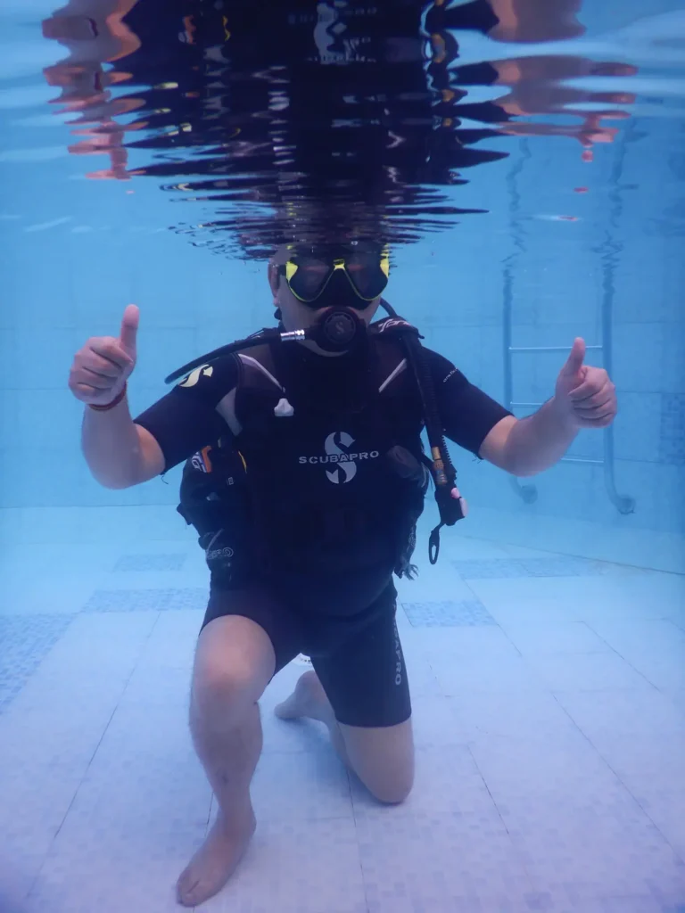 Diver kneeling on the pool floor gives two thumbs up, his reflection mirrored in the surface above