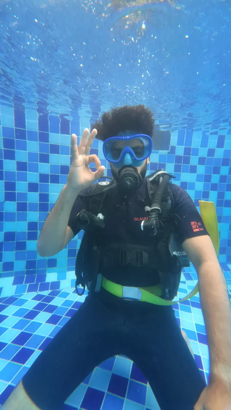 Diver in a blue mask makes an OK sign while kneeling above blue mosaic pool tiles
