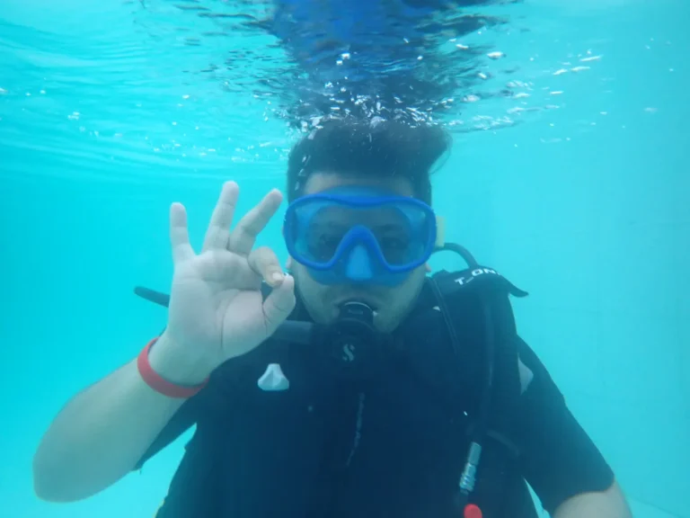 Diver in a blue mask makes an OK sign just below the bright turquoise surface of the pool