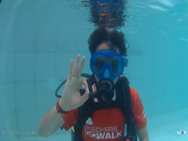 A diver in a blue mask and orange shirt kneels underwater by the pool tiles, giving an OK sign