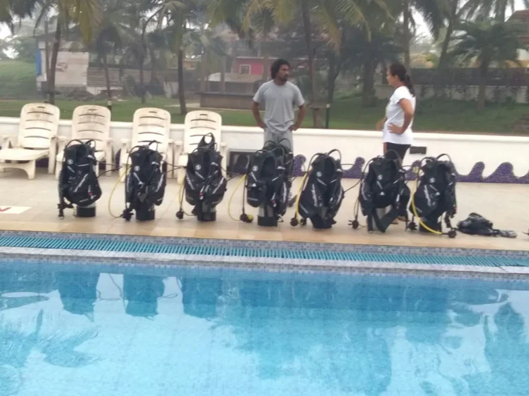 Row of assembled scuba kits stands along the pool edge while two staff members talk nearby