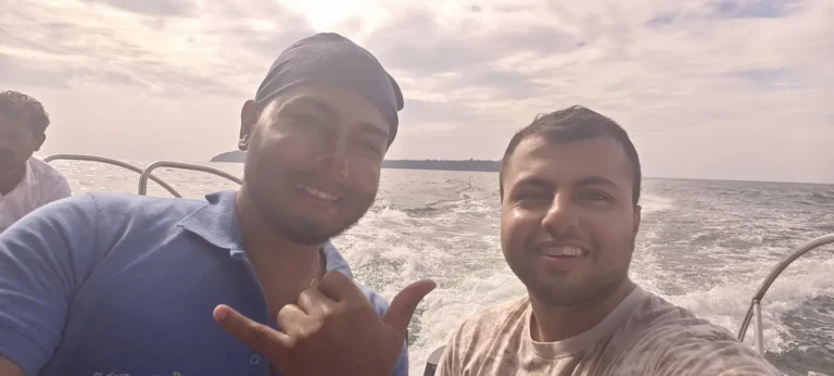 Two men smile and throw a shaka on a speeding boat, with white wake and cloudy sky behind