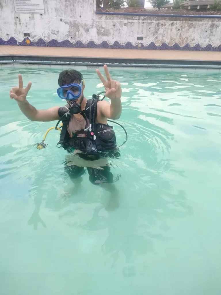 Student in a blue mask and BCD stands waist deep in the pool, flashing peace signs with both hands