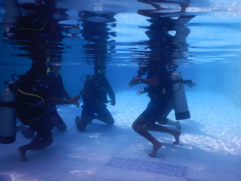Underwater view of students in scuba gear kneeling on a pool floor beneath a rippling surface