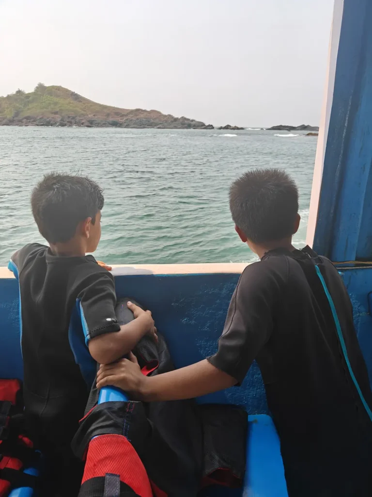 Two boys in wetsuits stand at the side of a boat looking out toward a rocky green island