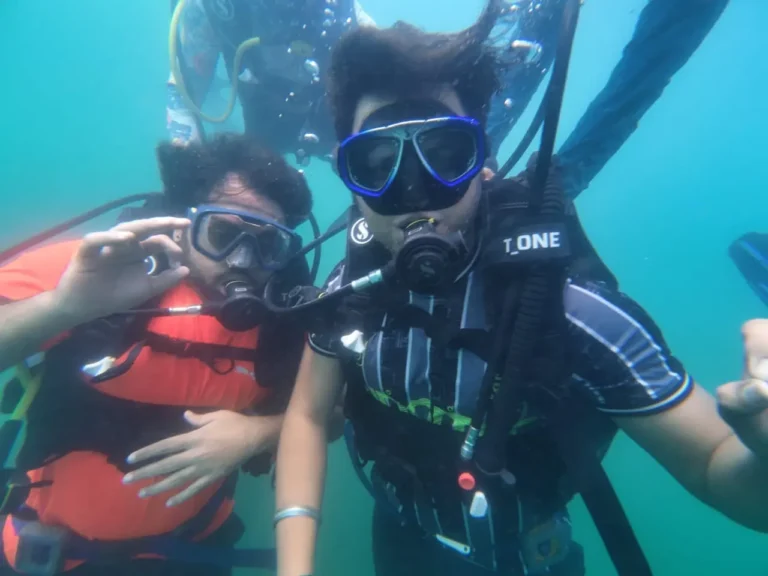 Two divers lean in close for a selfie in green-blue water, one making an OK sign