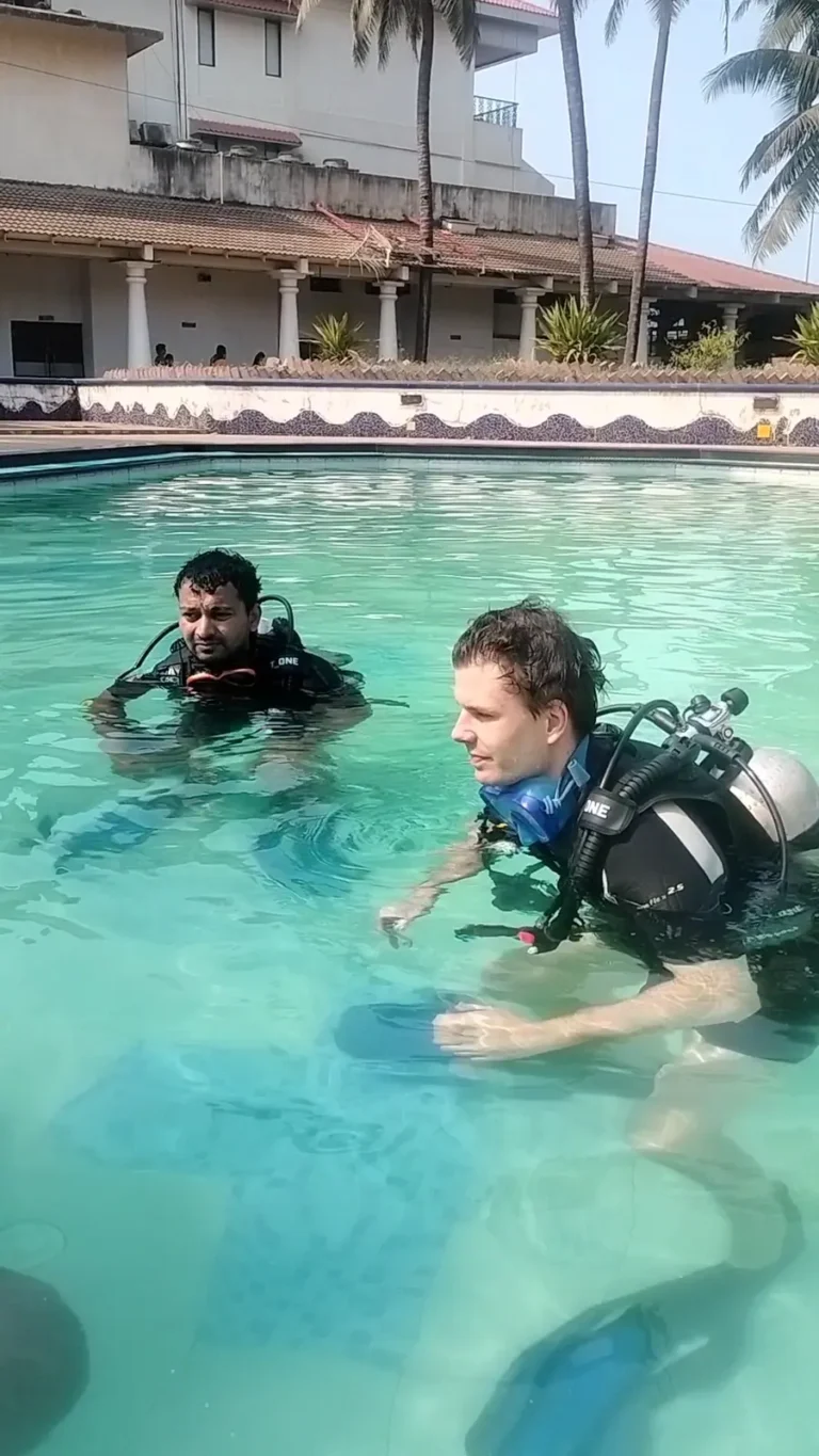 Two divers in BCDs float at the surface of a resort pool with palm trees and a building behind
