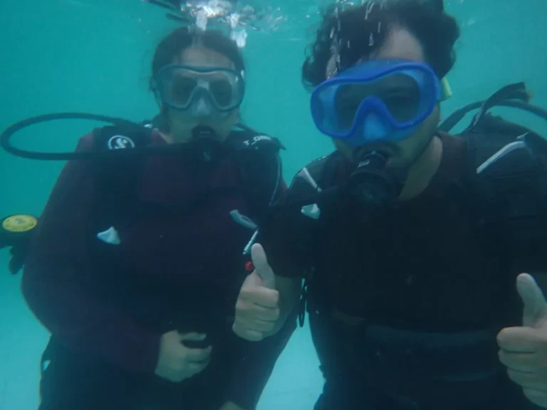 Two divers side by side give thumbs up in blue-green water, bubbles rising above them