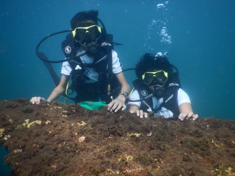 Two young divers in yellow masks peek over a rock underwater, bubbles rising beside them