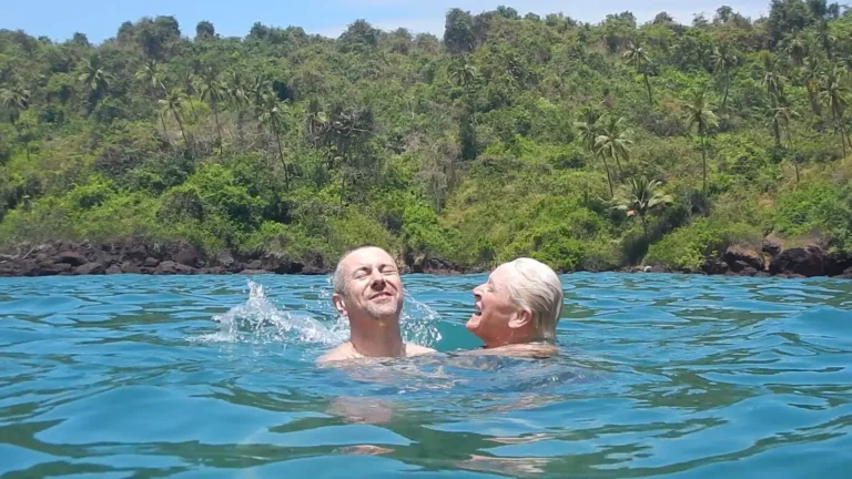 Two men laugh and splash in clear blue sea water below a hillside thick with palms and trees