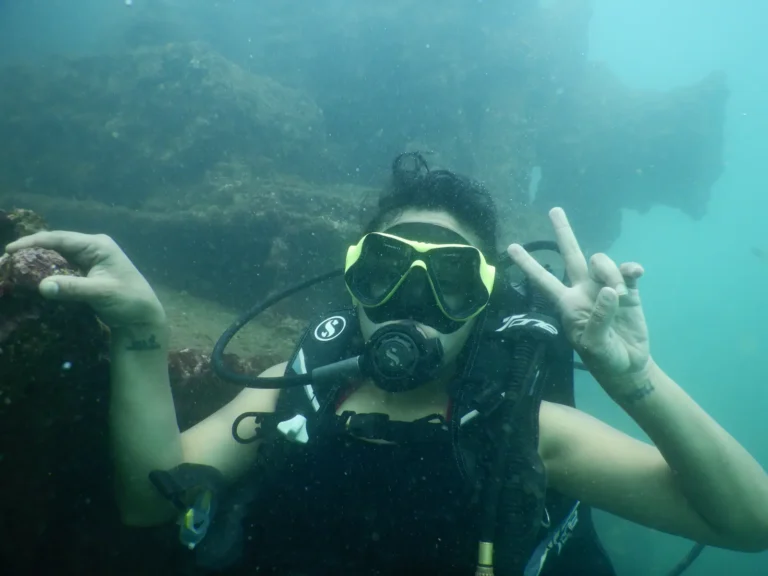 Woman diver in a yellow mask flashes a peace sign while holding onto a rock in green water