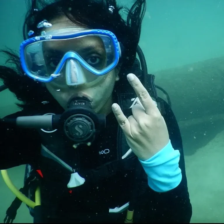 Woman diver in a blue mask makes a rock on hand sign, with dark hair floating around her face