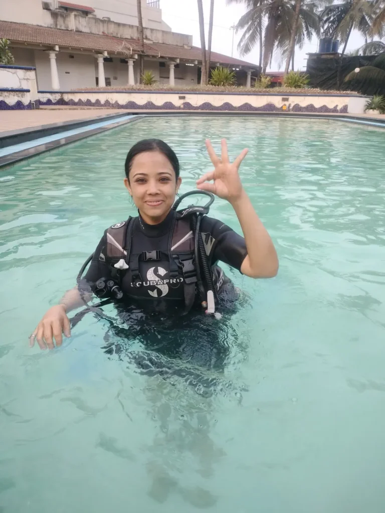 Smiling woman in a black BCD stands in the resort pool and makes an OK sign with one hand