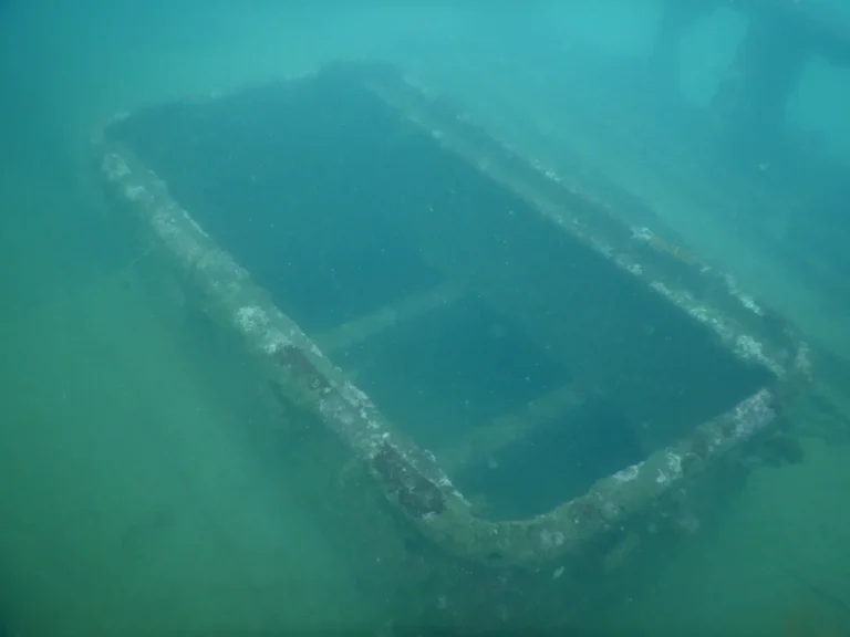 A rectangular hatch frame on a sunken wreck, crusted with growth, seen from above in green water