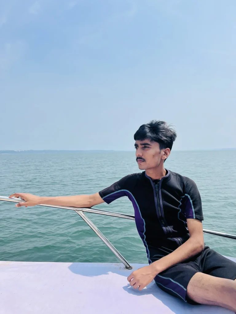 Young man in a black shorty wetsuit leaning on the railing of a boat deck, looking out to sea